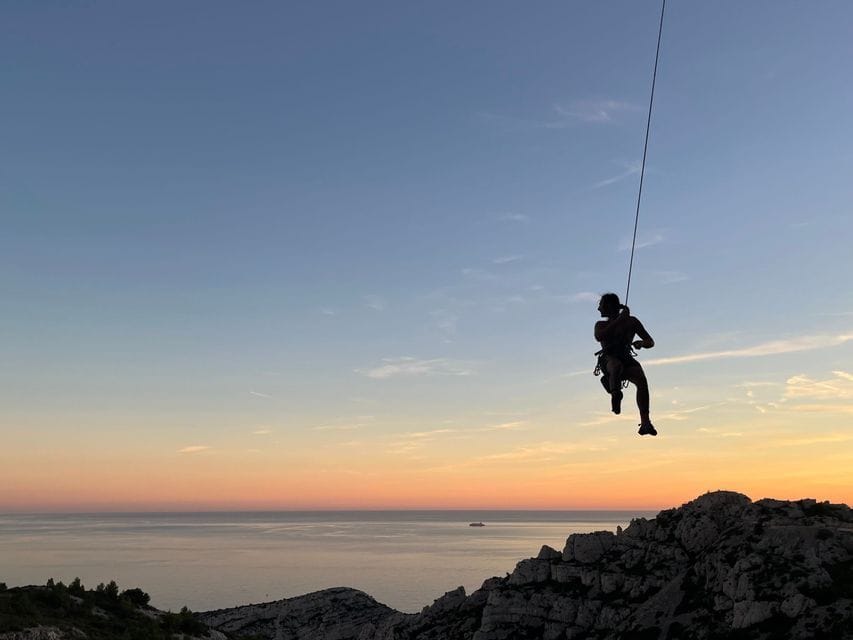 Silhouette d'une personne en rappel le long d'une falaise rocheuse surplombant la mer au coucher du soleil.