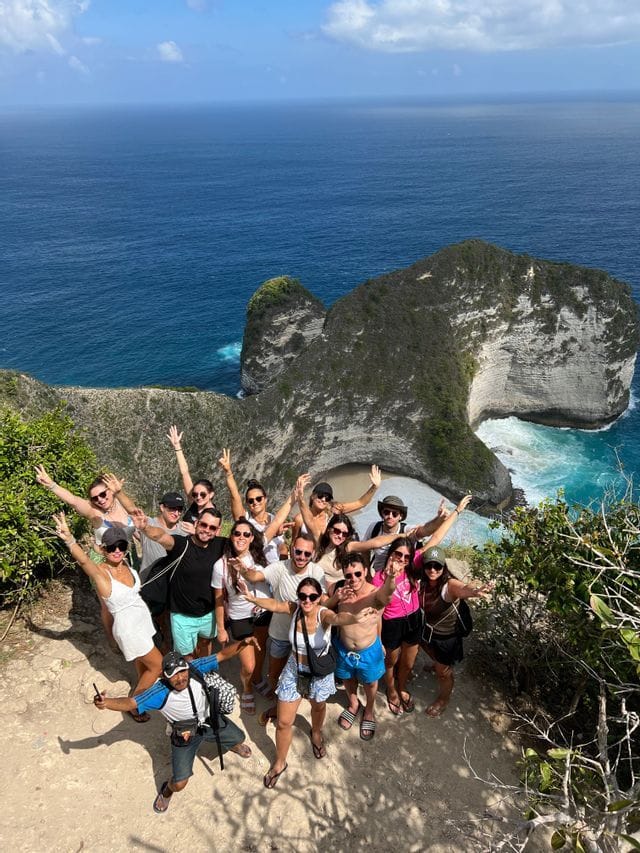 Un gruppo WeRoad sorride e posa su un punto panoramico sulla scogliera, con una formazione rocciosa drammatica e l'oceano turchese sottostante.