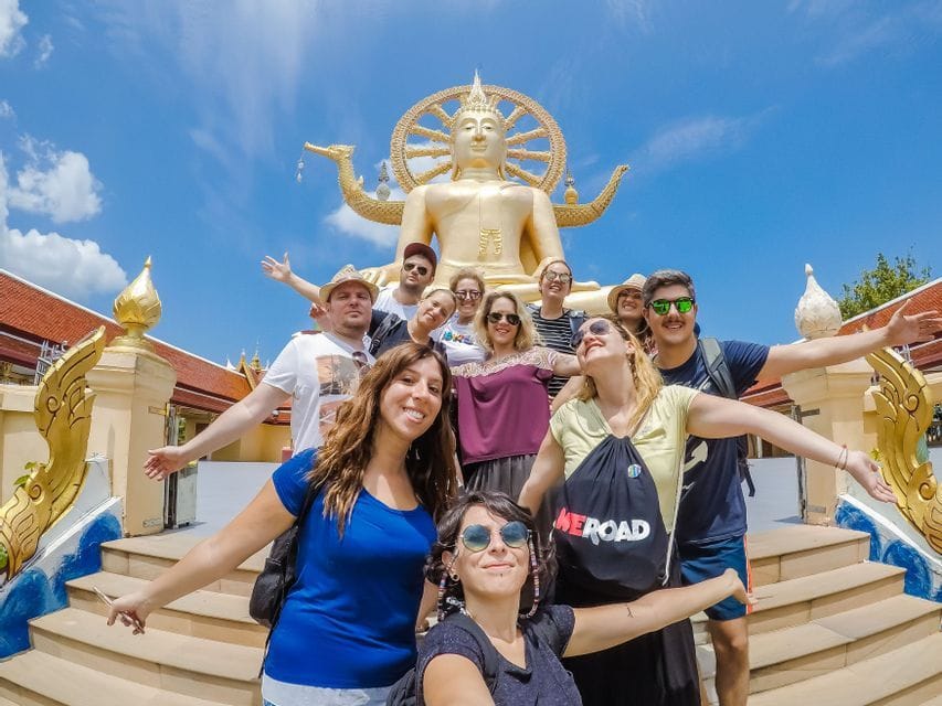 Un groupe WeRoad en voyage posant pour une photo sur les marches d'un temple, devant une grande statue de Bouddha dorée, sous un ciel bleu.