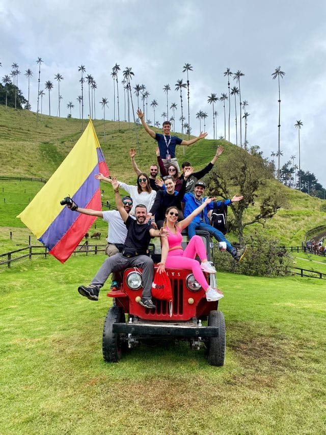 Un groupe WeRoad en voyage pose sur un 4x4 rouge arborant un drapeau colombien, dans une vallée de grands palmiers.