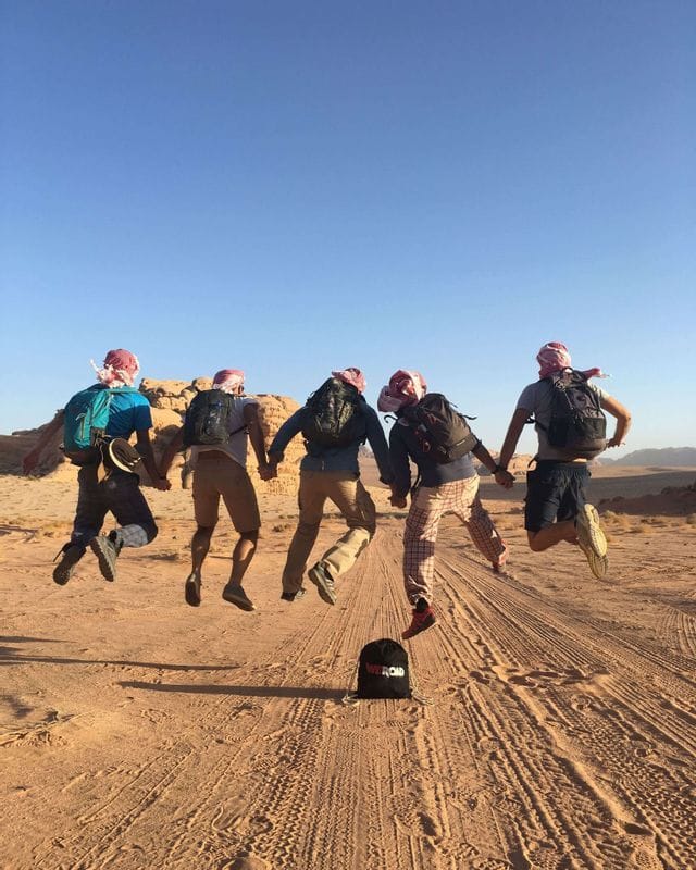 Five people on a WeRoad group trip hold hands and jump in the air in a desert landscape, seen from behind.