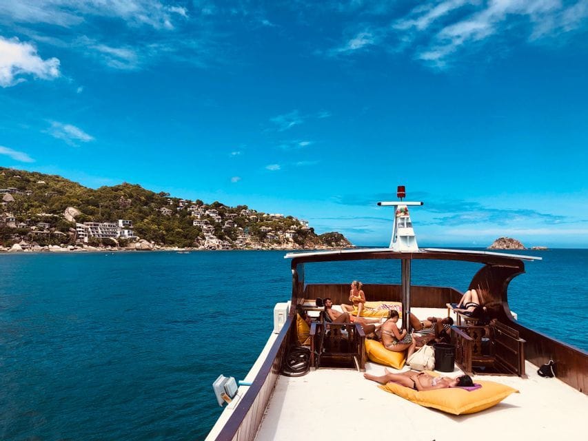 Un groupe WeRoad se détend et prend le soleil sur le pont d'un bateau, naviguant sur la mer bleue le long d'une île verte et vallonnée.