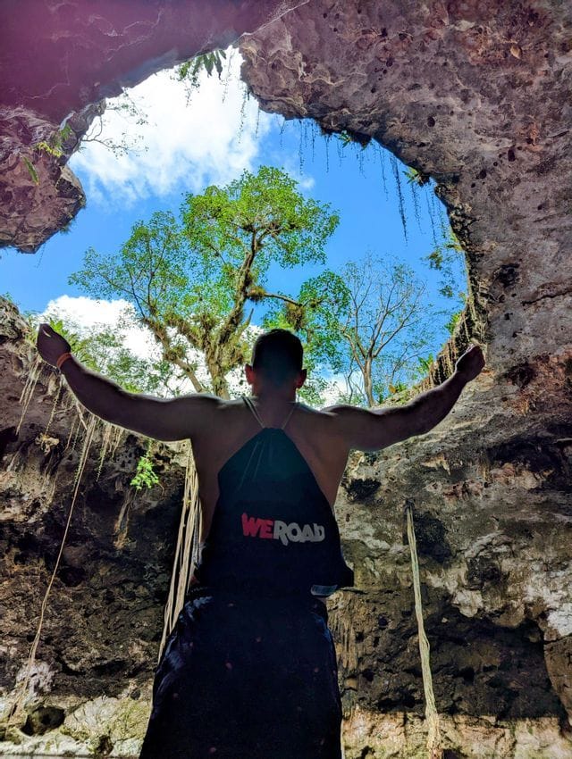 Un homme de dos portant un sac à dos WeRoad lève les bras à l'intérieur d'une grotte, regardant le ciel et les arbres à travers une ouverture.