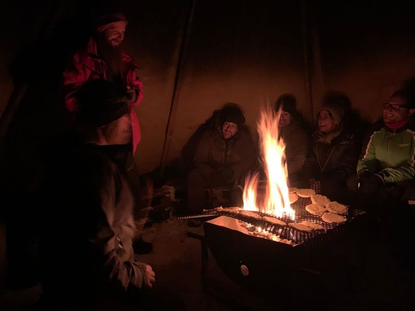 A WeRoad group trip gathers around a bonfire at night, cooking food on a grill inside a dark tent.