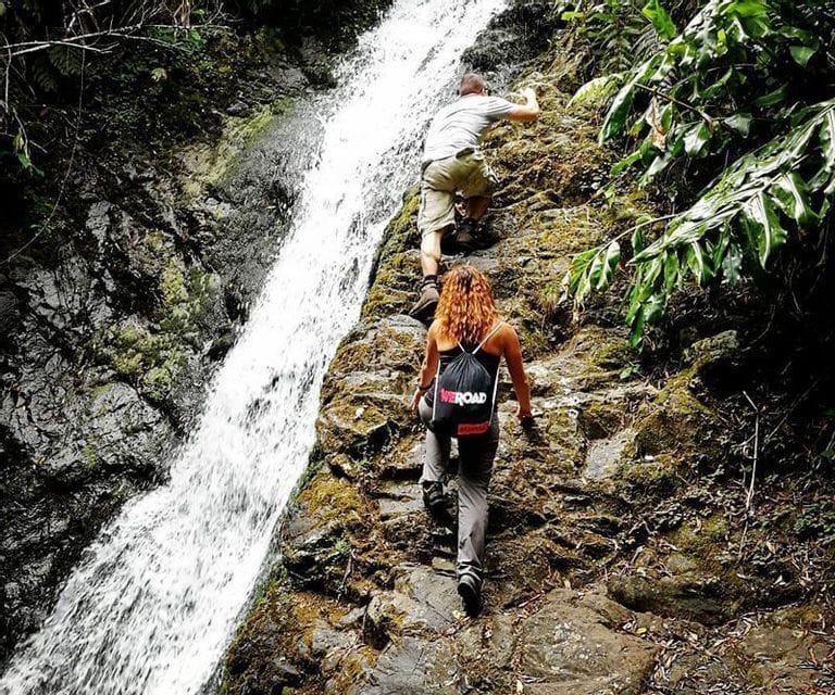 A man and a woman from a WeRoad group trip climbing up a steep, mossy rock face next to a cascading waterfall.