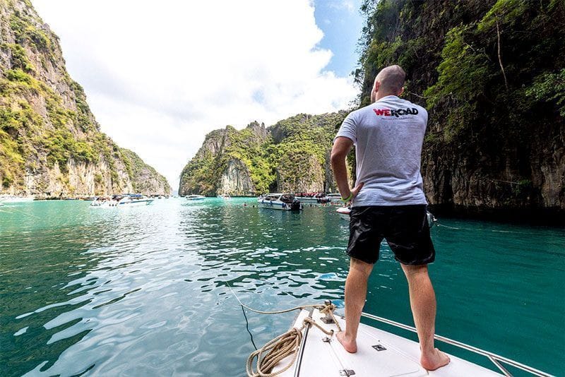 A man in a WeRoad t-shirt stands on the bow of a boat, looking at a tropical lagoon surrounded by steep cliffs.