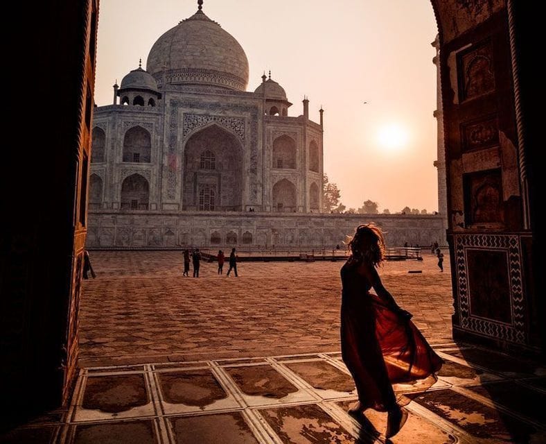 Une femme en robe rouge se tient en silhouette dans une arche de pierre, regardant le Taj Mahal au lever du soleil.