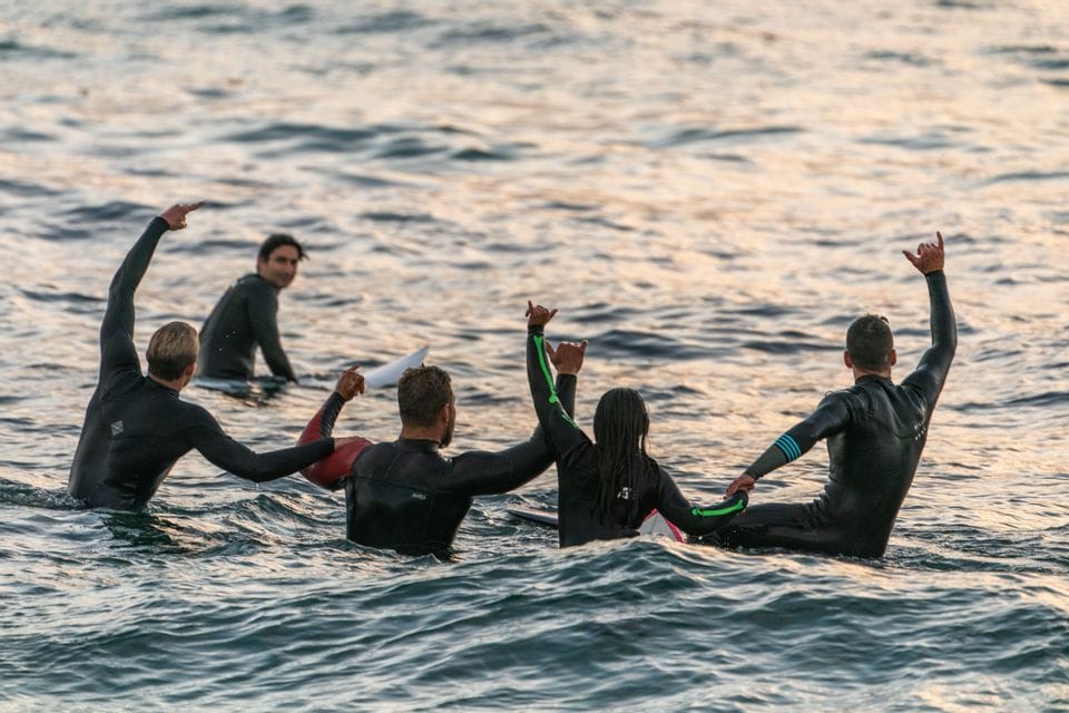 Eine WeRoad-Gruppe von Surfern feiert bei Sonnenuntergang auf ihren Boards im Meer, von hinten gesehen.