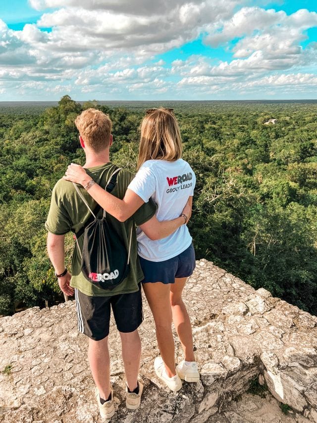Ein Mann und eine Frau einer WeRoad-Reisegruppe stehen auf einer Steinplattform und blicken über einen weiten grünen Dschungel.