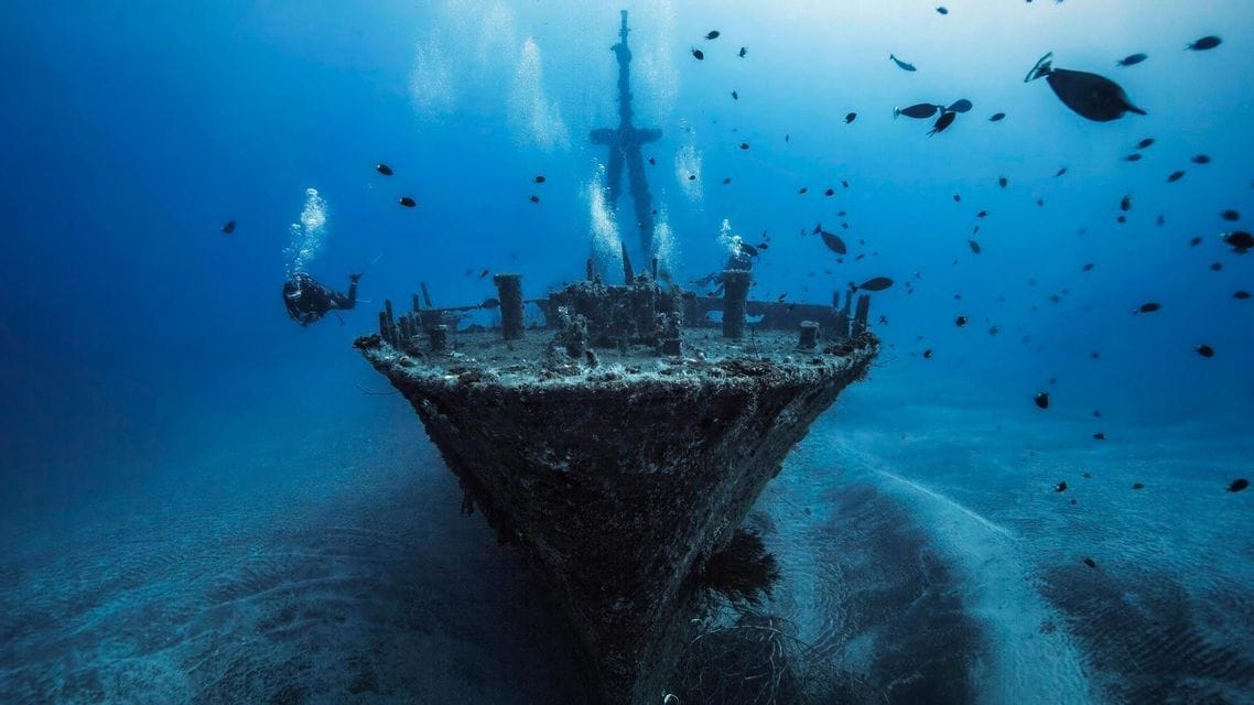 Un gruppo WeRoad di subacquei esplora un grande relitto adagiato sul fondale sabbioso, circondato da pesci.