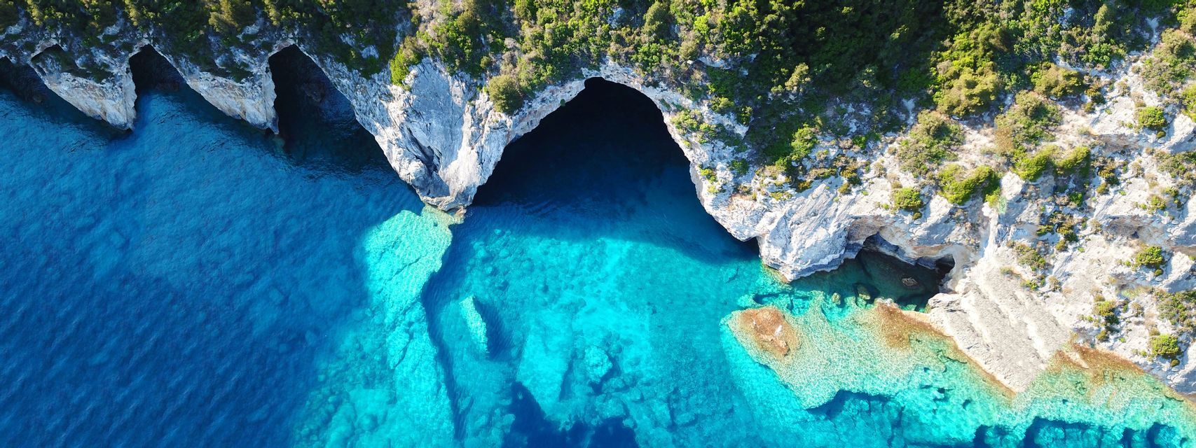 Vista aérea de cuevas marinas en un acantilado rocoso blanco con vegetación verde, rodeadas de aguas turquesas y azul profundo.