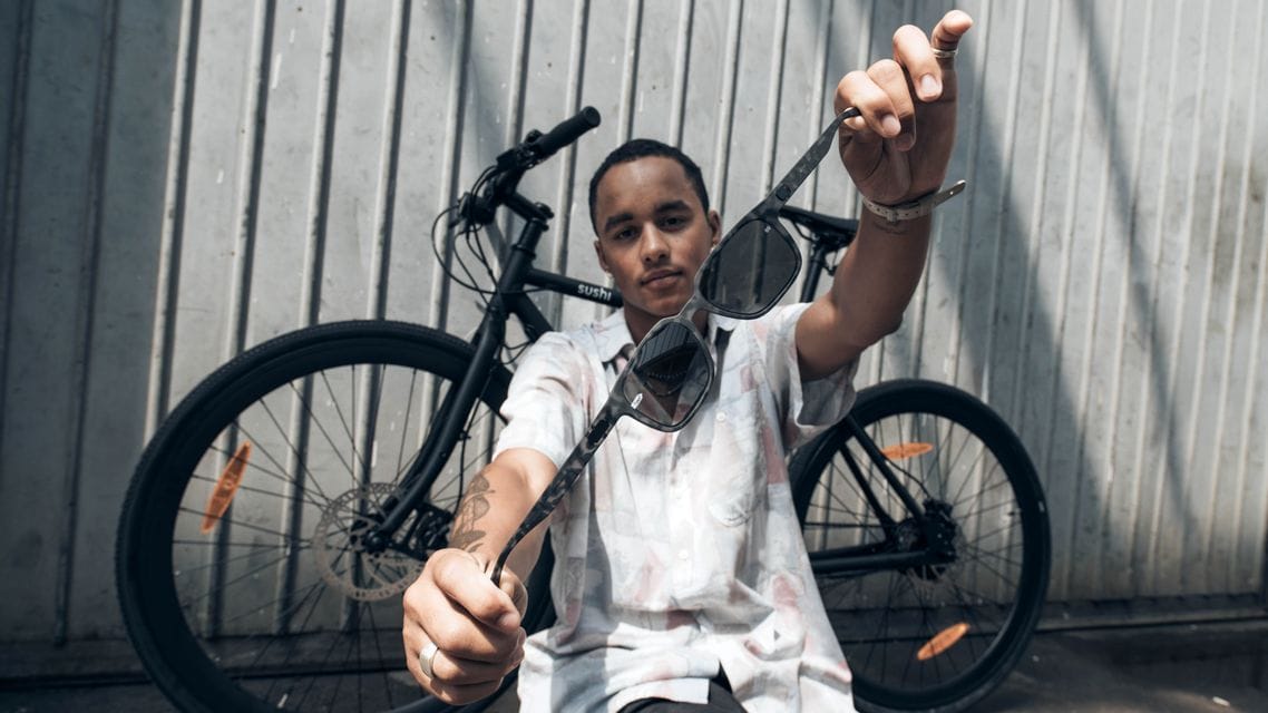 Ein Mann hält eine Sonnenbrille in die Kamera, mit einem schwarzen Fahrrad hinter sich vor einer Wellblechwand.
