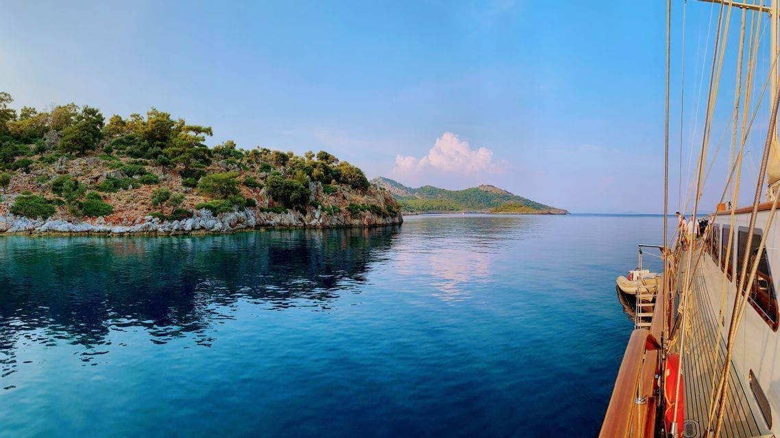 Vista dal ponte in legno di una barca che naviga su acque calme e blu, lungo una costa rocciosa e verde, sotto un cielo sereno.
