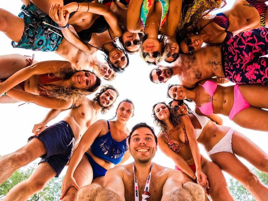 Una selfie en contrapicado de un grupo WeRoad sonriente en traje de baño, reunidos en círculo contra un cielo brillante.