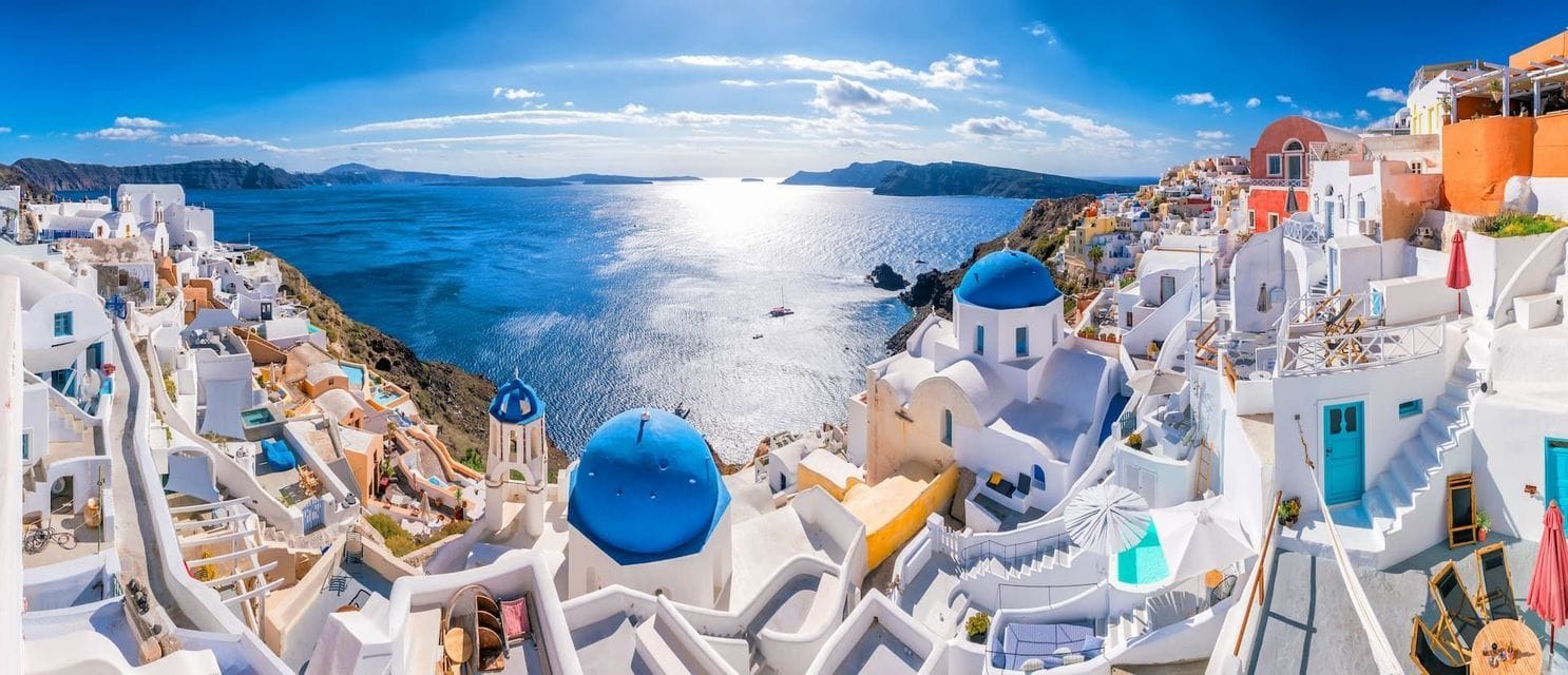 Una vista panorámica de edificios blancos e iglesias de cúpulas azules en un acantilado con vistas a un mar soleado bajo un cielo azul.