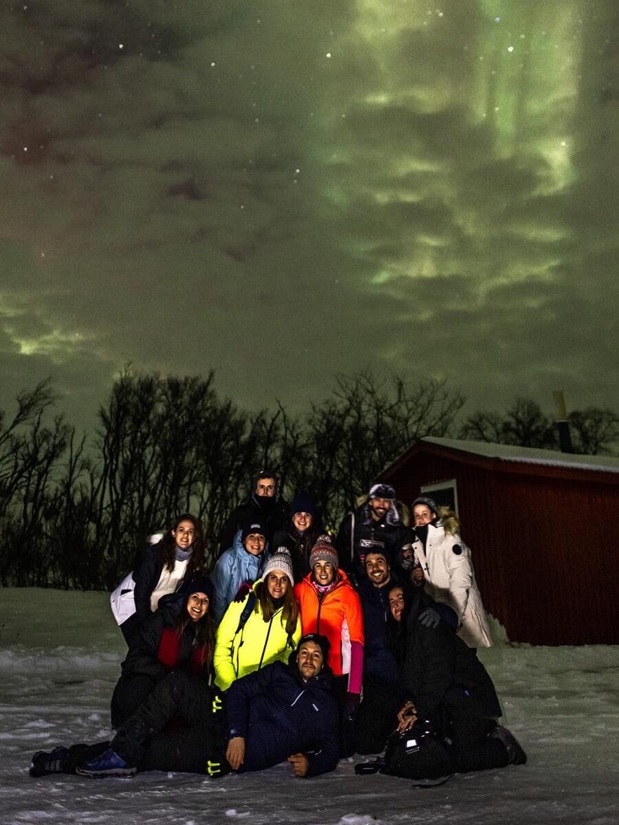 Un voyage de groupe WeRoad pose en vêtements d'hiver sur la neige sous des aurores boréales vertes, près d'un chalet rouge.