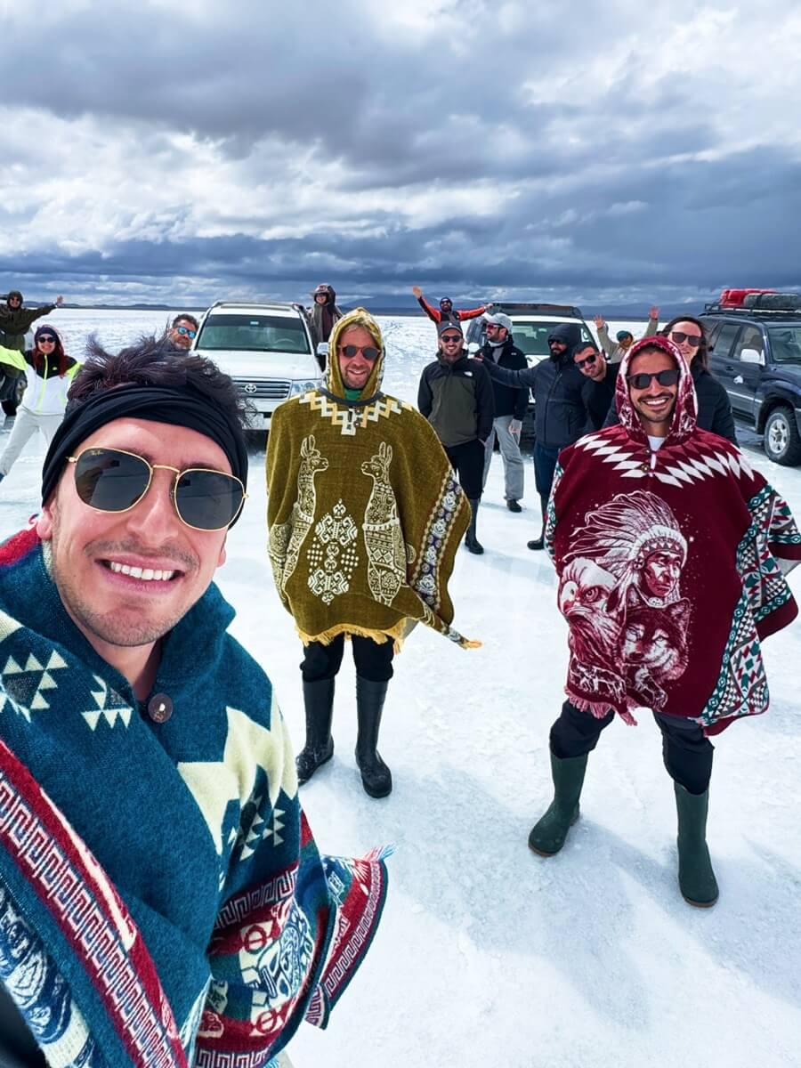A WeRoad group trip selfie of travelers wearing colorful ponchos on a salt flat with 4x4 vehicles behind.