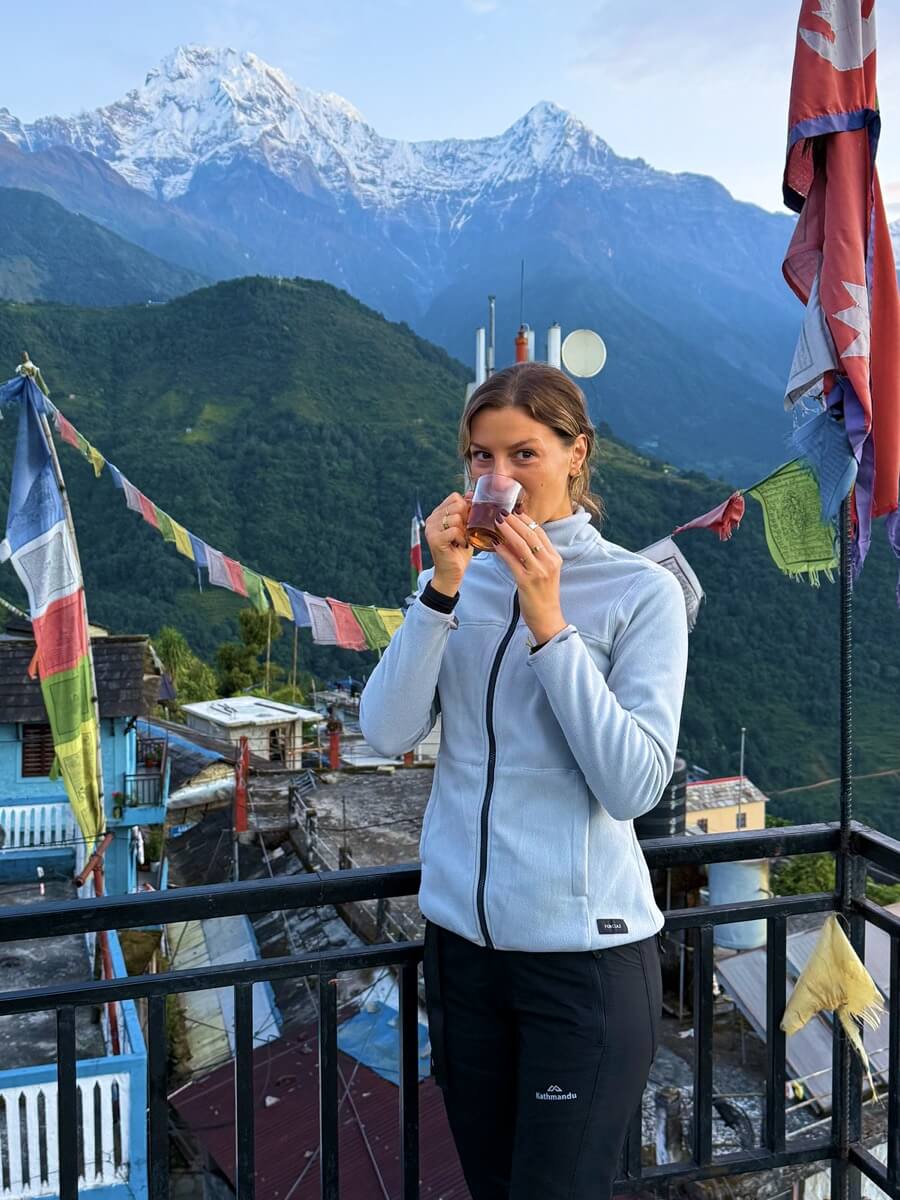 Woman in a fleece jacket sips from a glass on a balcony with colorful prayer flags and snow-capped mountains behind her.