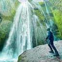 Persona su una sporgenza rocciosa di fronte a una cascata che scende lungo una scogliera coperta di muschio verde.