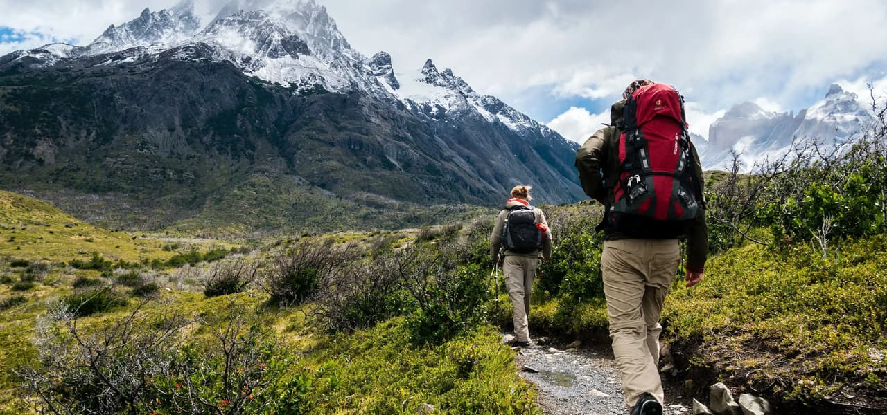 Zwei Wanderer mit großen Rucksäcken wandern auf einem schmalen Pfad durch ein grünes Tal in Richtung zerklüfteter, schneebedeckter Berge.