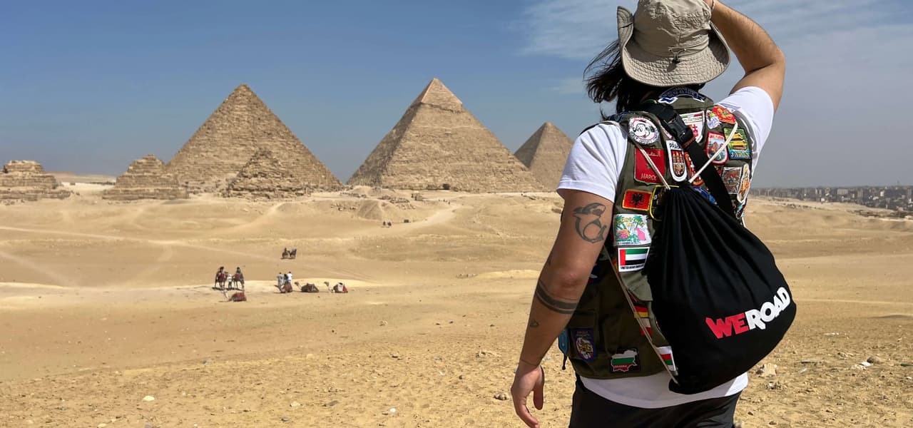 Un viaggiatore di spalle, con un cappello e uno zaino WeRoad, ammira grandi piramidi nel deserto.