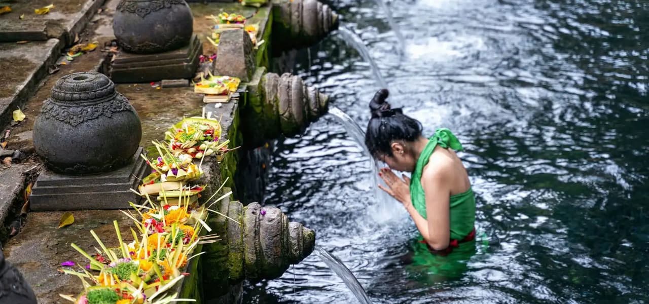 Una mujer con un top verde realiza un ritual de purificación, inclinando su cabeza bajo un chorro de agua de una fuente de piedra.