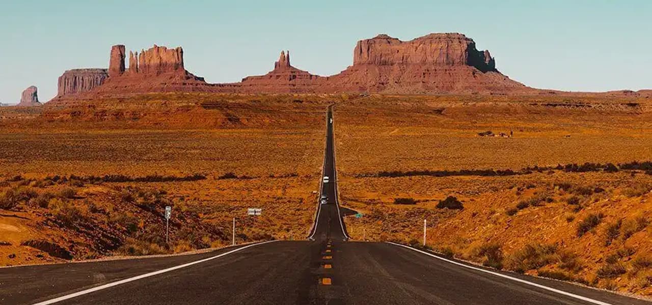 Una larga y recta carretera se extiende por un paisaje desértico hacia imponentes formaciones rocosas rojas bajo un cielo azul claro.