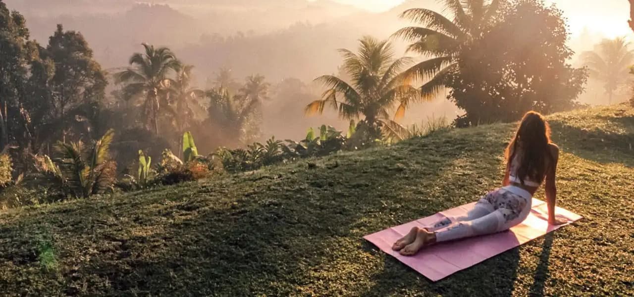 Une femme en posture de yoga sur un tapis, regardant une vallée verdoyante et brumeuse avec des palmiers au lever du soleil doré.