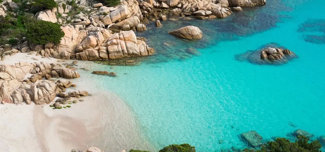 Cala de aguas turquesas y arena blanca rodeada de rocas de granito y vegetación mediterránea.
