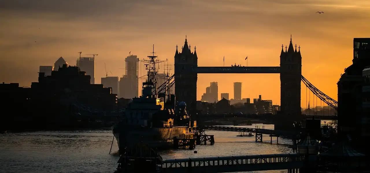 Silhouetten einer Zugbrücke, eines Kriegsschiffs und einer Stadtkulisse vor einem goldenen Sonnenuntergangshimmel über einem breiten Fluss.