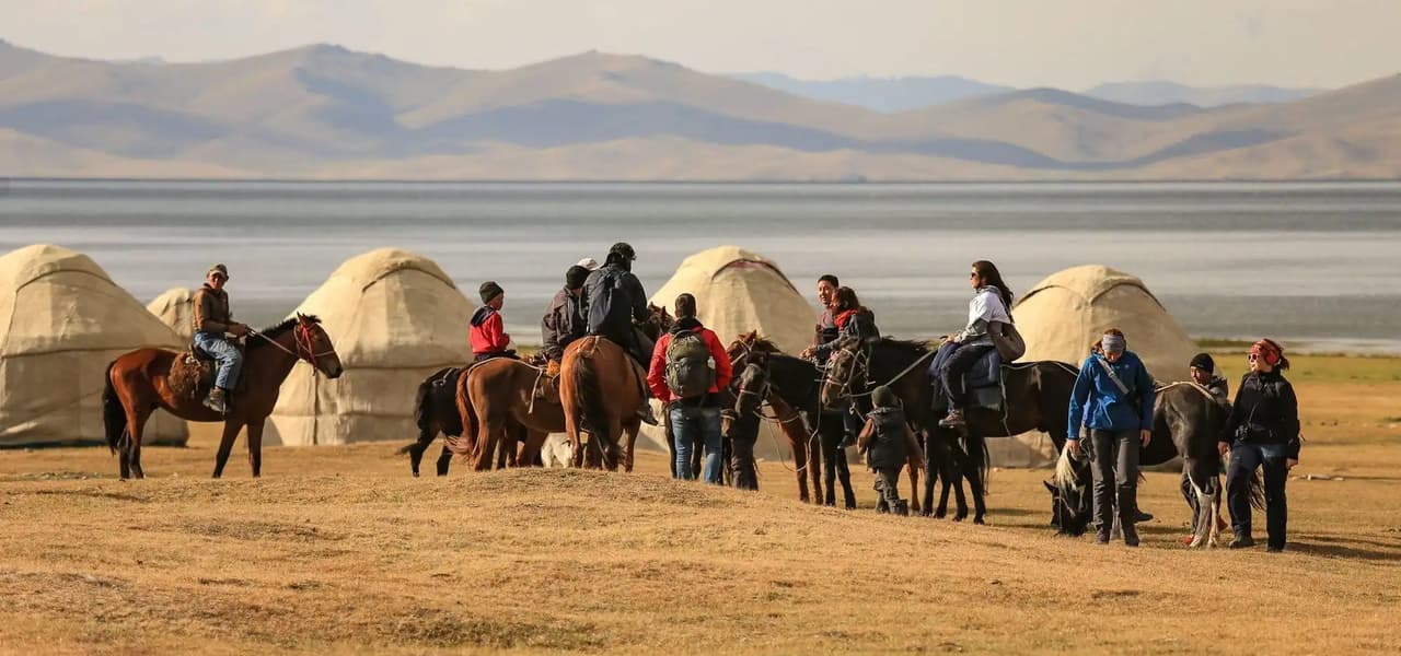 Un grupo de viajeros a caballo junto a un campamento de yurtas a orillas de un lago entre montañas.
