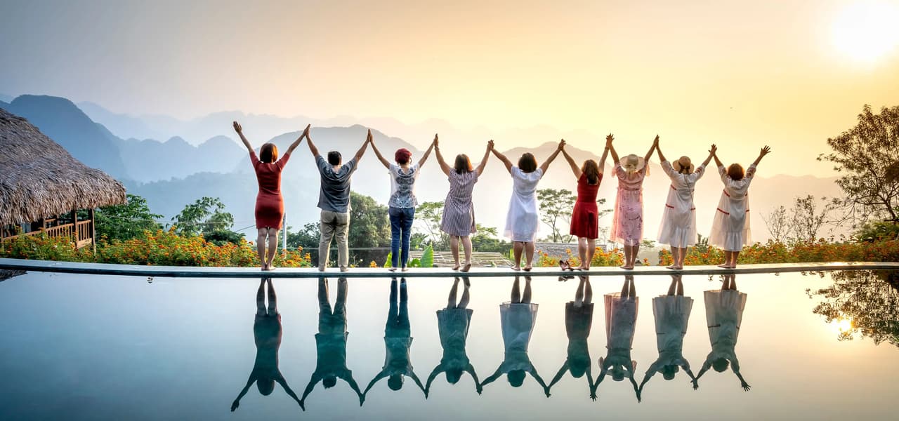 Un voyage de groupe WeRoad, main dans la main et bras levés au bord d'une piscine à débordement surplombant les montagnes, qui se reflètent dans l'eau.