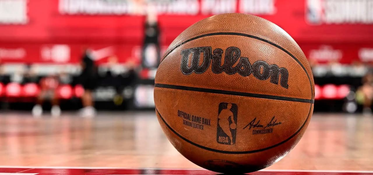Un gros plan d'un ballon de basketball officiel Wilson NBA posé sur un parquet lustré avec un arrière-plan rouge.