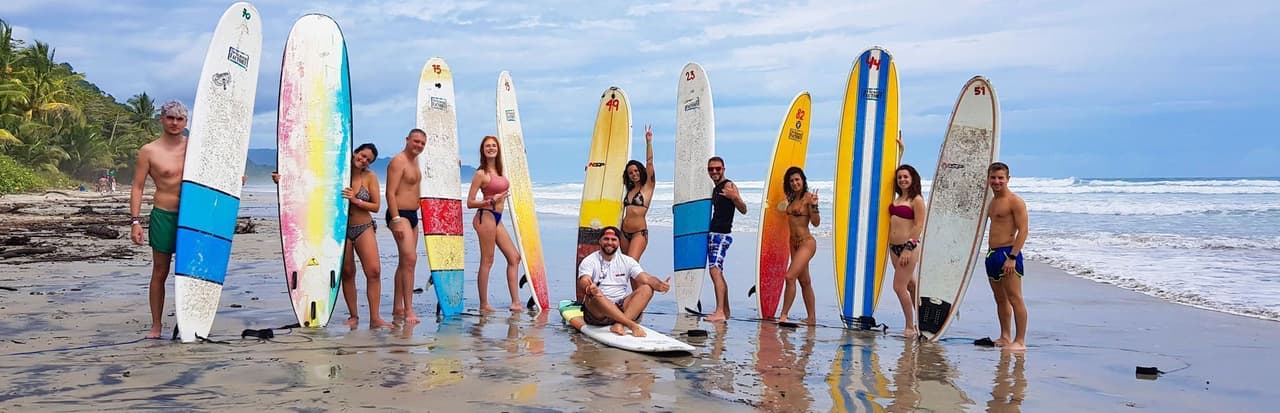 Un voyage en groupe WeRoad posant avec des planches de surf colorées sur une plage de sable humide avec l'océan en arrière-plan.