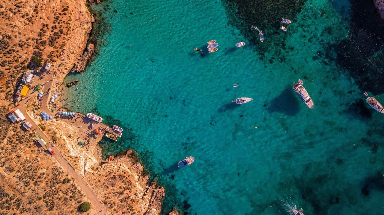 Vue aérienne de nombreux bateaux flottant dans une eau turquoise limpide le long d'une côte rocheuse avec une petite plage bondée.