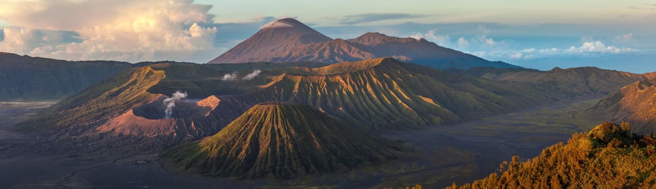 Ein Panoramablick auf eine Vulkanlandschaft mit mehreren Kratern, von denen einer Rauch ausstößt, und einem großen Berggipfel im Hintergrund.