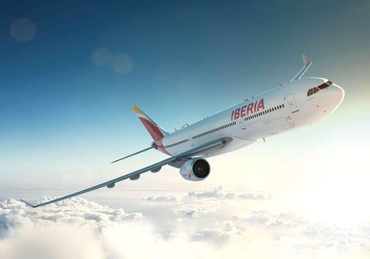 Un avión de Iberia sobrevuela un extenso paisaje urbano con dos rascacielos prominentes durante un atardecer brumoso.
