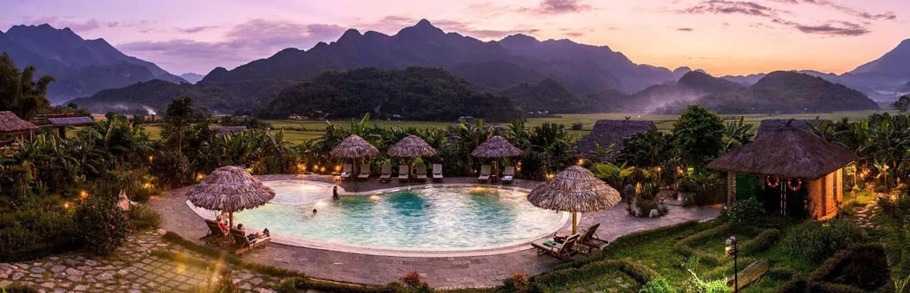 Un edificio tradizionale in legno accanto a una piscina a sfioro, che riflette il cielo e le nuvole al tramonto, con montagne sullo sfondo.