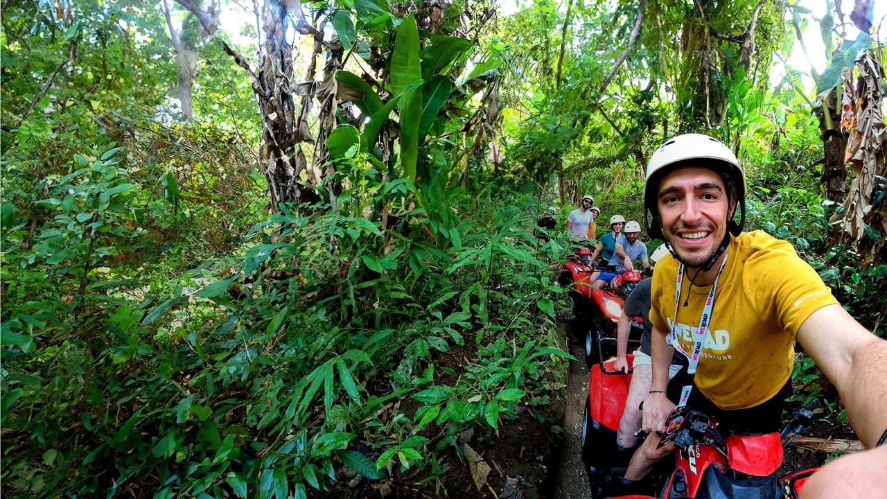 A WeRoad group trip on red quad bikes riding in a line on a narrow trail through a dense green jungle.