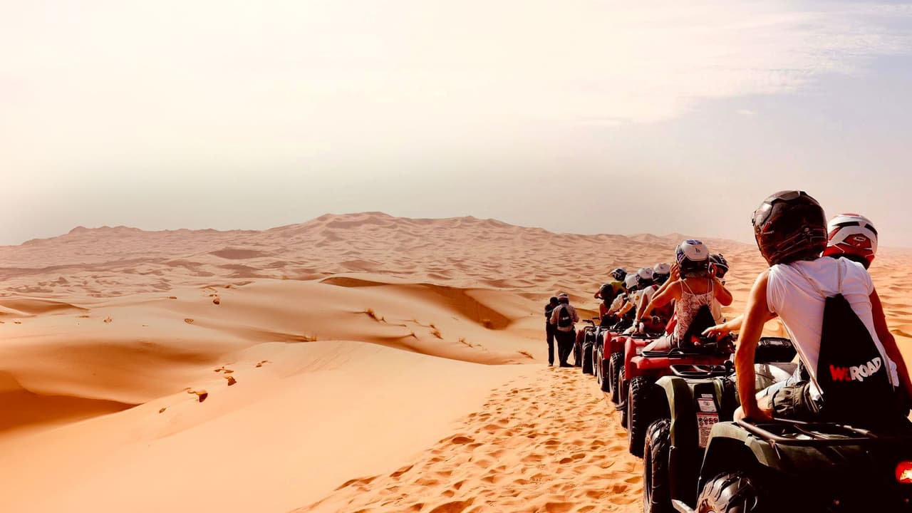 Un groupe WeRoad en voyage, casqué, est assis sur des quads alignés sur des dunes de sable du désert sous un ciel lumineux.