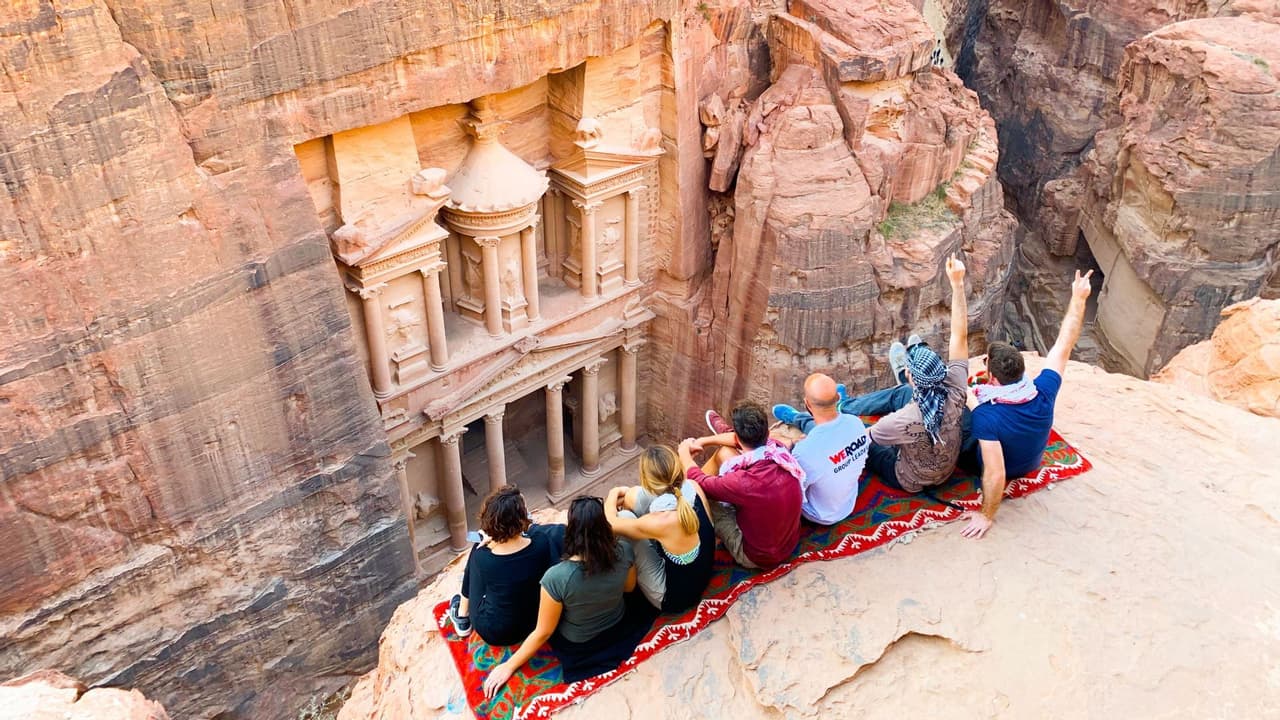 Un grupo de WeRoad se sienta sobre una alfombra en un saliente de acantilado, contemplando un gran templo tallado en un cañón de roca roja.