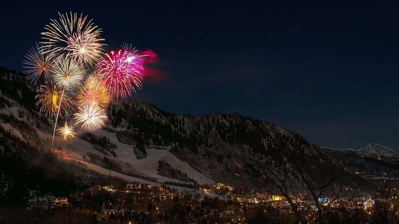 Fuochi d'artificio colorati esplodono nel cielo notturno sopra una città ai piedi di una montagna innevata.