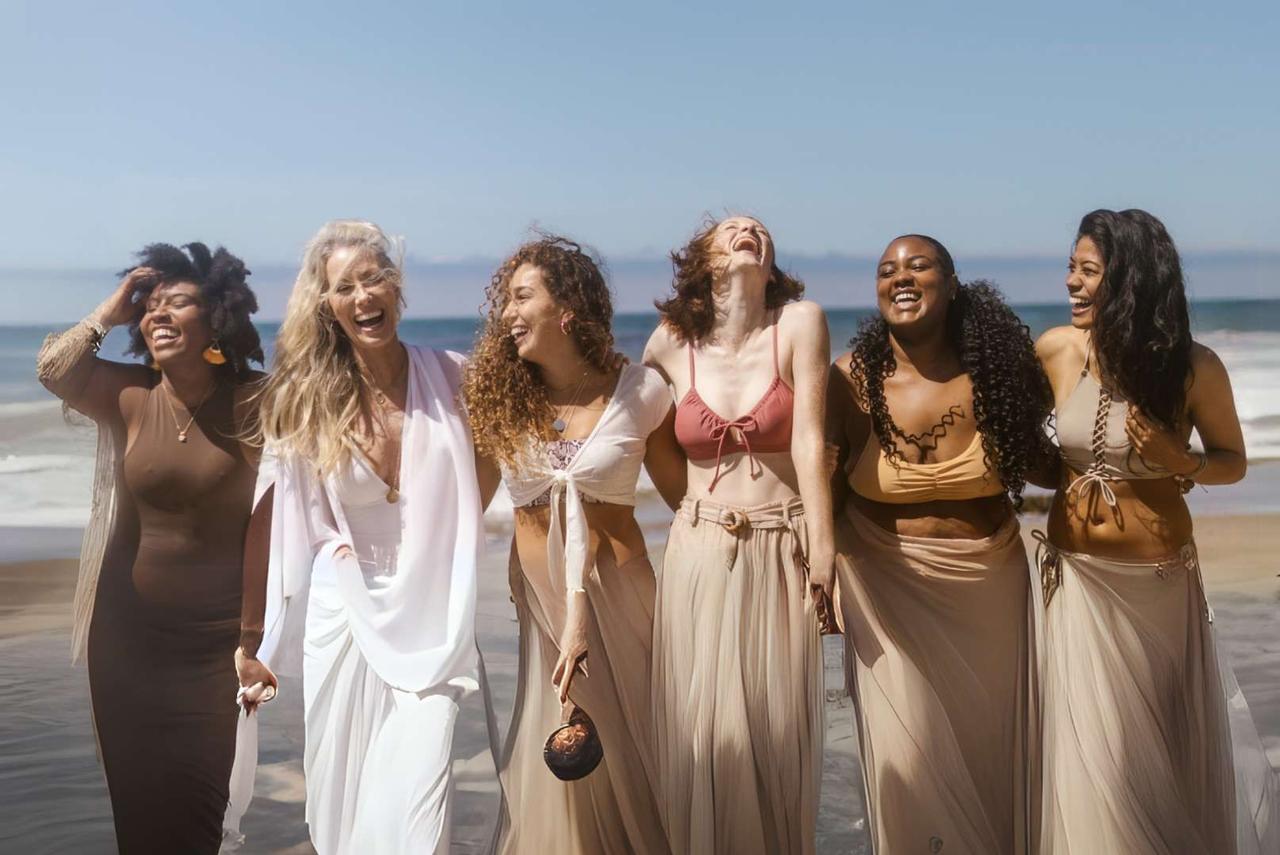 Un groupe WeRoad de six femmes riant ensemble sur une plage de sable, avec l'océan en arrière-plan.