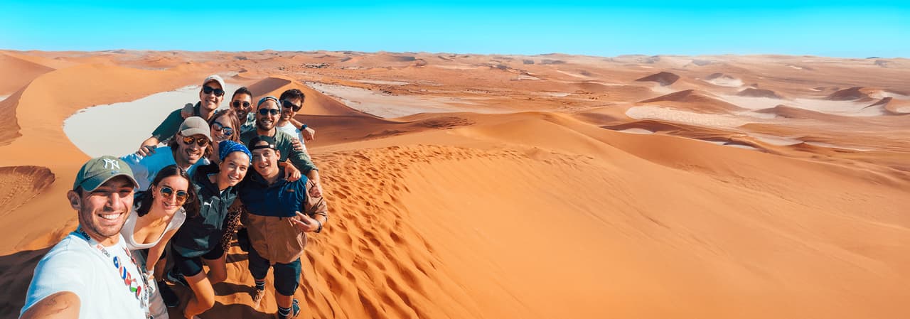 Un viaggio di gruppo WeRoad scatta un selfie su una grande duna di sabbia arancione nel deserto sotto un cielo azzurro e limpido.