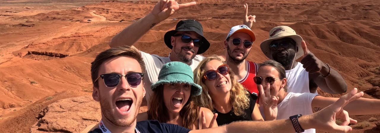 Un voyage de groupe WeRoad prend un selfie souriant devant des formations rocheuses rouges sous un ciel partiellement nuageux.
