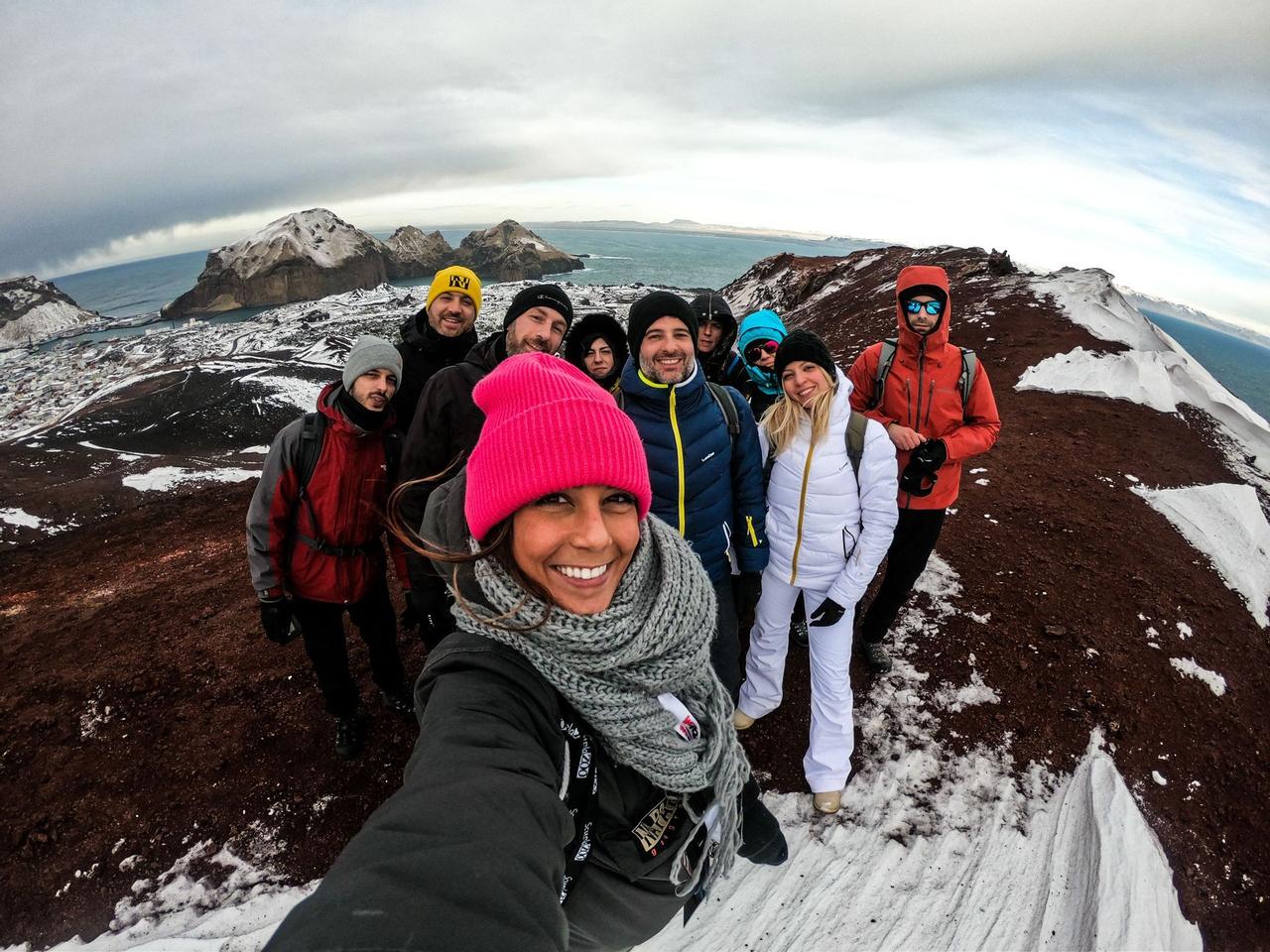 Un viaje en grupo de WeRoad sonríe para una selfie en una cresta de montaña nevada, con islas y el mar de fondo.