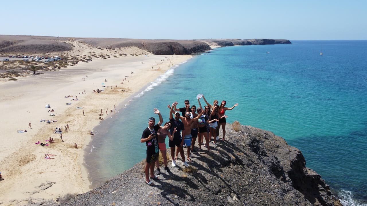Un grupo de WeRoad posa y sonríe en un acantilado rocoso con vistas a una playa de arena y un océano turquesa.