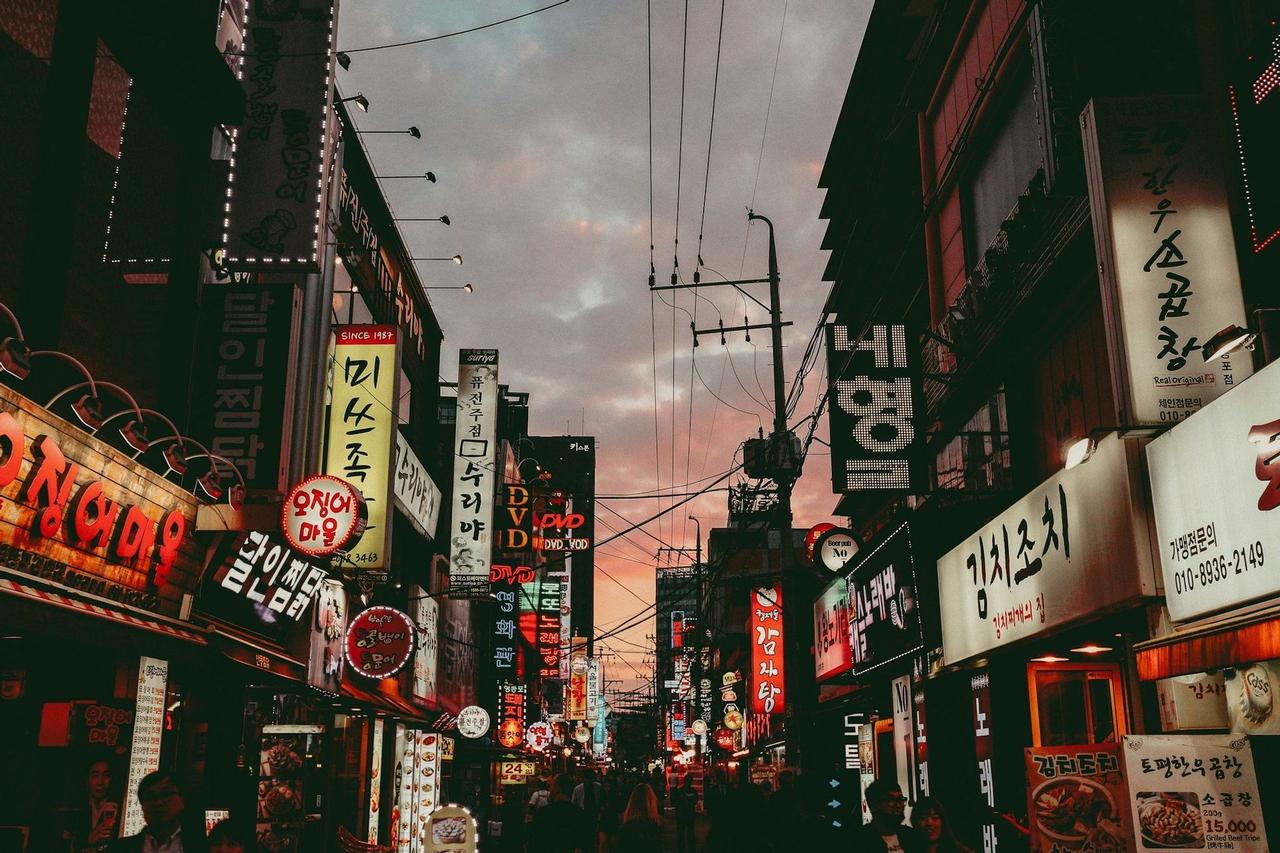 Una calle estrecha de la ciudad vista desde abajo, bordeada de edificios cubiertos de letreros luminosos con letras coreanas bajo un cielo crepuscular.