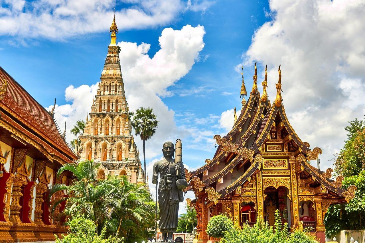 Ein kunstvoll verzierter goldener Tempel und eine hohe steinerne Stupa stehen neben einer Mönchsstatue, umgeben von Grün unter blauem Himmel.