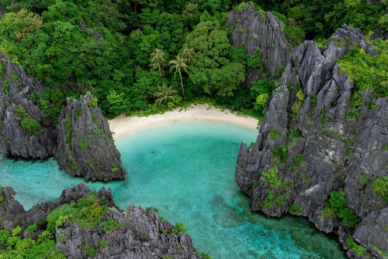 Vue aérienne d'une crique isolée avec une plage de sable blanc, entourée d'eaux turquoise, de formations rocheuses déchiquetées et d'une forêt verdoyante.